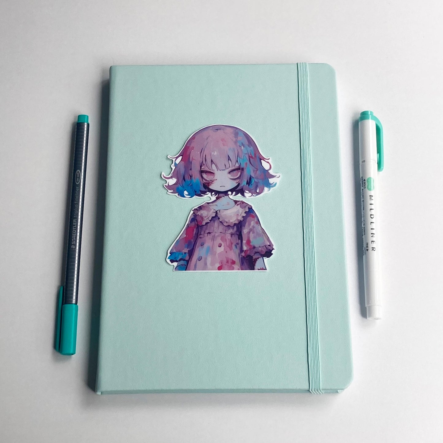 A sticker of an anime character with purple and blue hair in a ruffled dress is placed on a closed light teal notebook. A pen with a teal cap is on the left, and a teal marker is on the right.