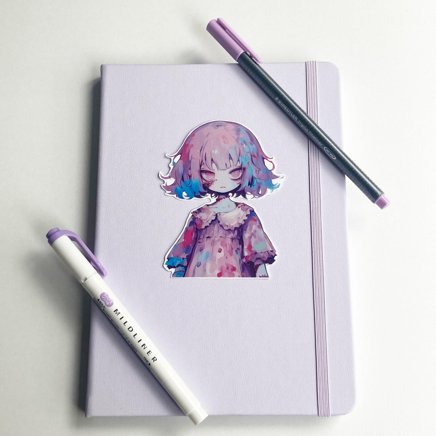A sticker of an anime character with purple and blue hair in a ruffled dress is placed on a closed light purple notebook. A purple marker and a pen with a purple cap lie diagonally across the notebook.