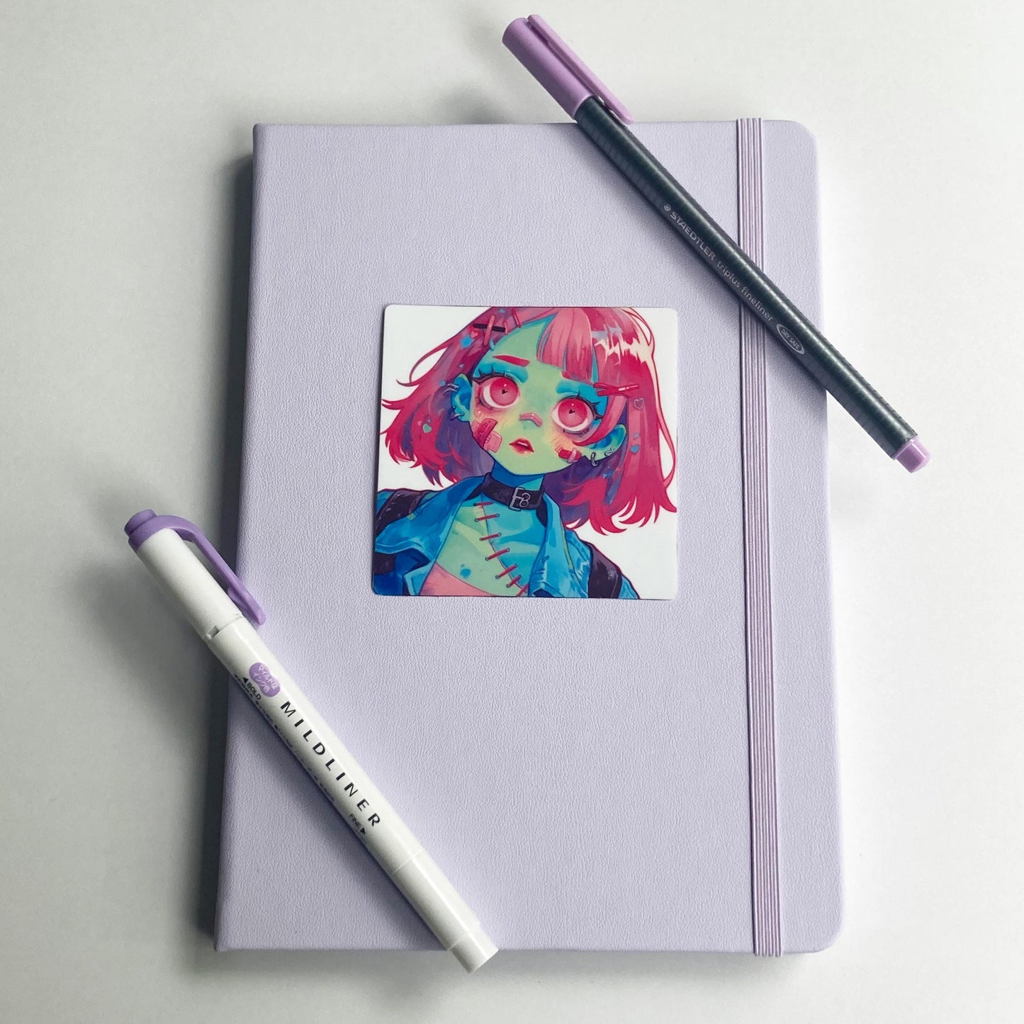 A lavender notebook with a sticker of an illustrated character with pink hair, green skin, and stitched details. A purple-tipped Mildliner marker and a gray fineliner pen with a purple cap are placed diagonally on the notebook.