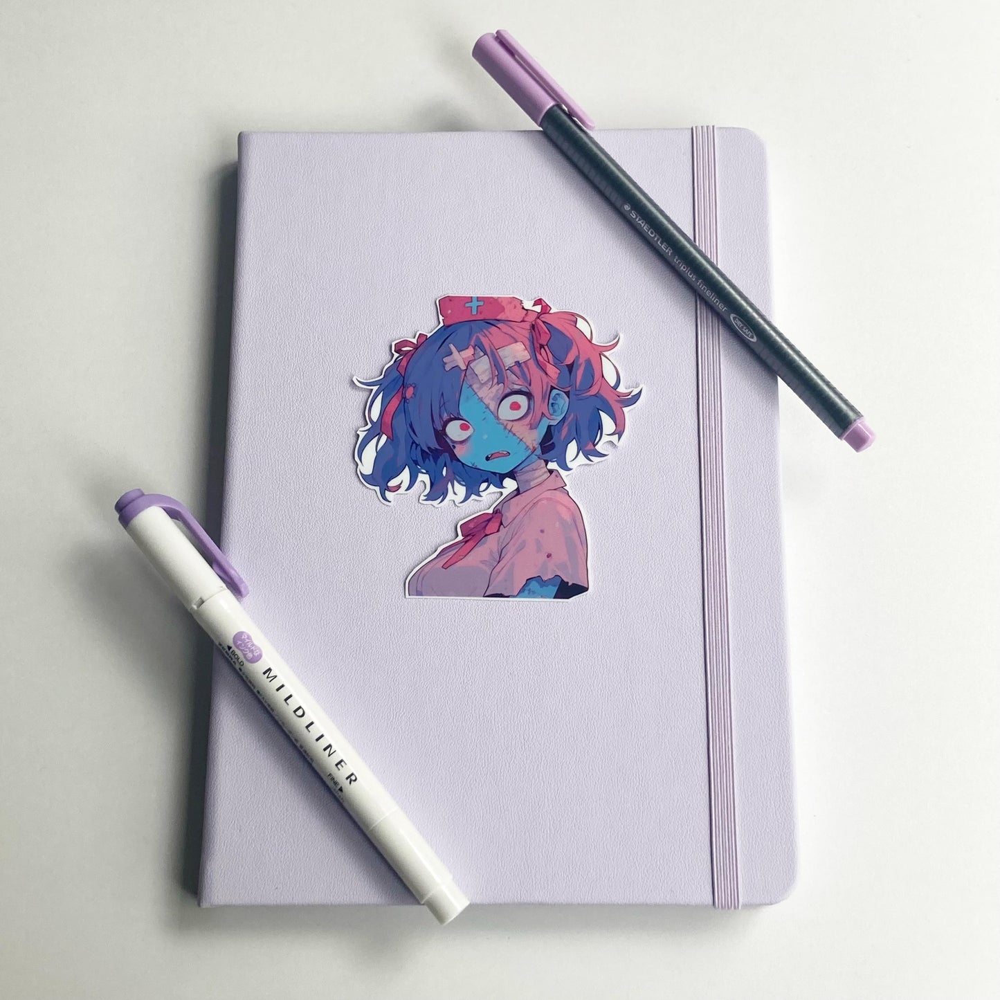 A blue and pink skinned zombie nurse sticker is on a lavender notebook. The notebook alsohas a highlighter and pen sitting on it. The sticker has purple and pink pigtails in bows. Wears a pink nurses outfit with tears and a nurse hat with cross