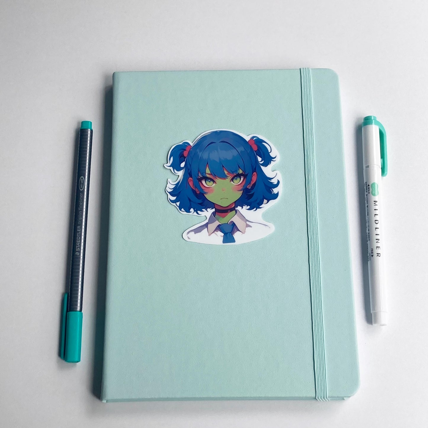 Sticker of an anime character with blue twin-tails, pink hair ties, green skin, and yellow eyes on a light teal notebook. The character wears a white shirt, blue tie, and black choker. A teal pen and matching highlighter are placed on either side