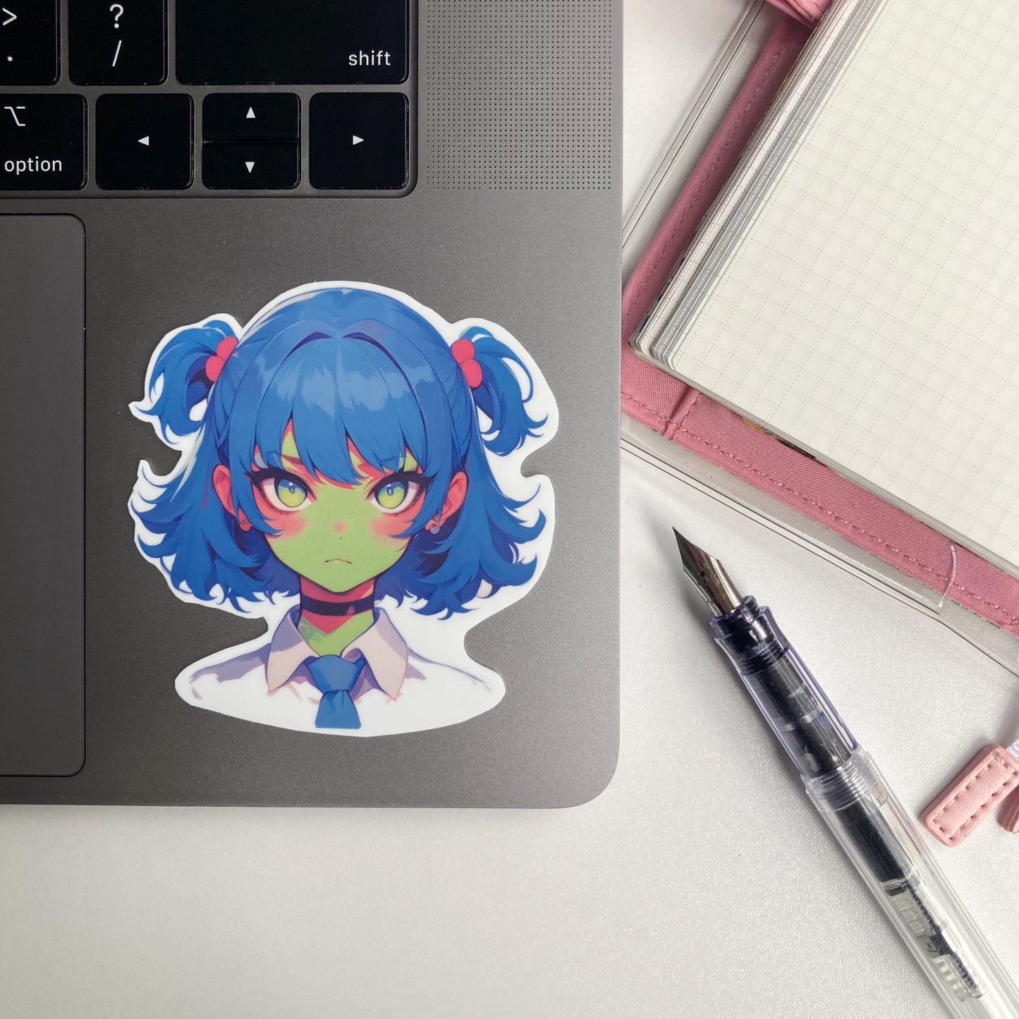 Sticker of an anime-style character with blue twin-tails, pink hair ties, green skin, and yellow eyes, wearing a white shirt, blue tie, and black choker. The sticker is placed on a laptop keyboard corner.