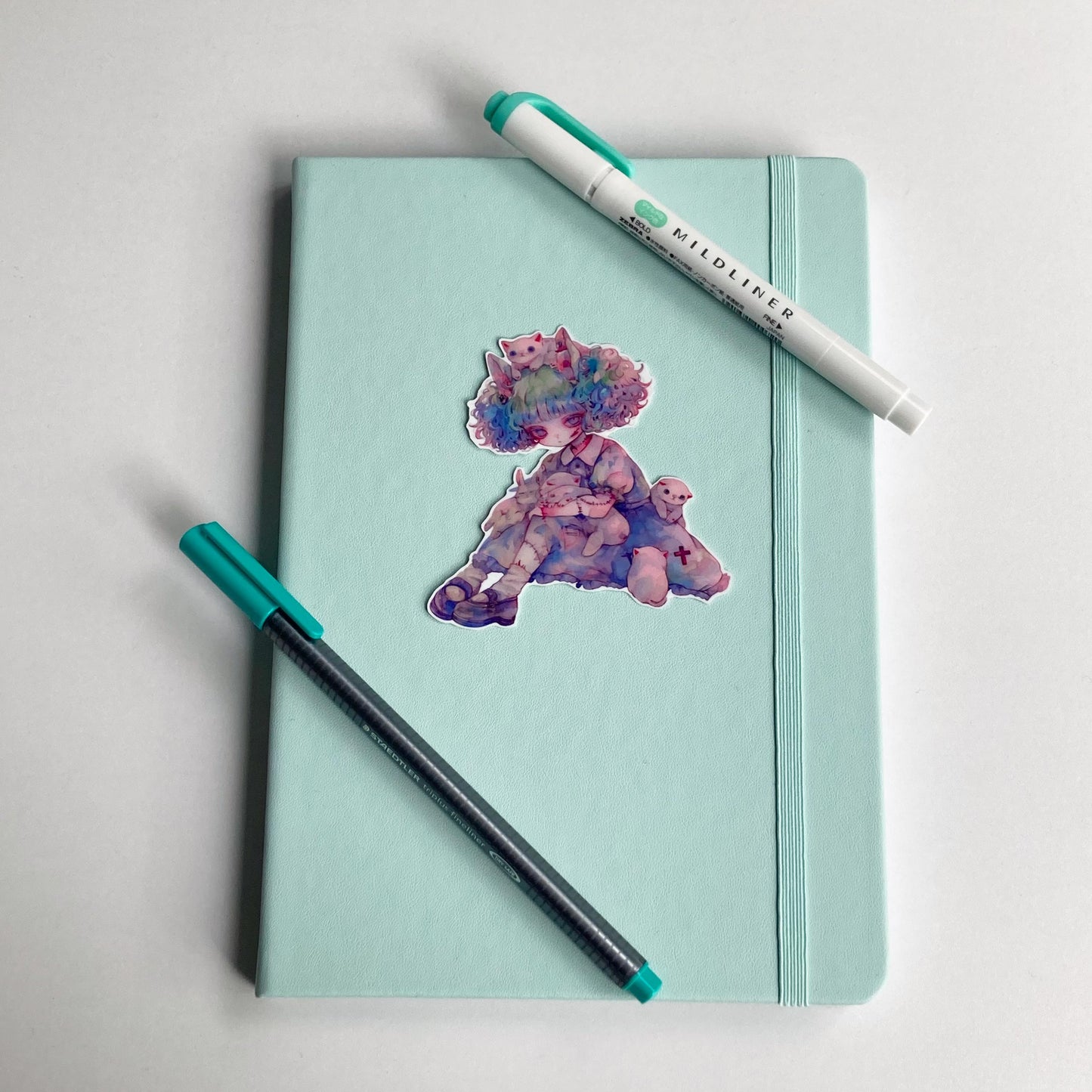 Sticker of an anime-style character with pastel curly hair, white cats, and bunnies, wearing a blue dress with stitched limbs. Sticker is on a light teal notebook with a teal pen and highlighter.