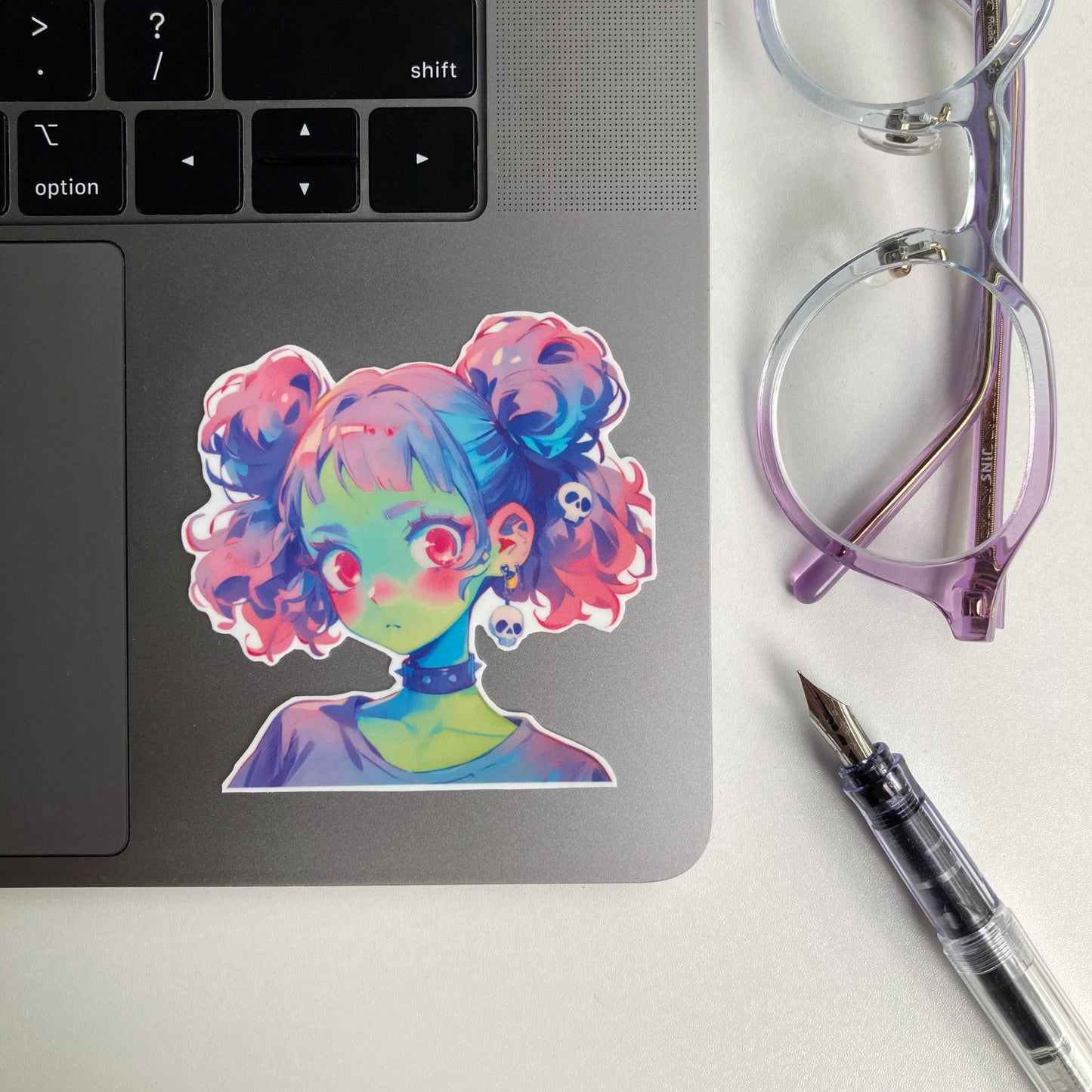 Sticker of an anime-style character with pastel pink and blue twin buns, green skin, and large pink eyes. The character wears a black choker and skull earrings. The sticker is placed on a laptop keyboard corner.
