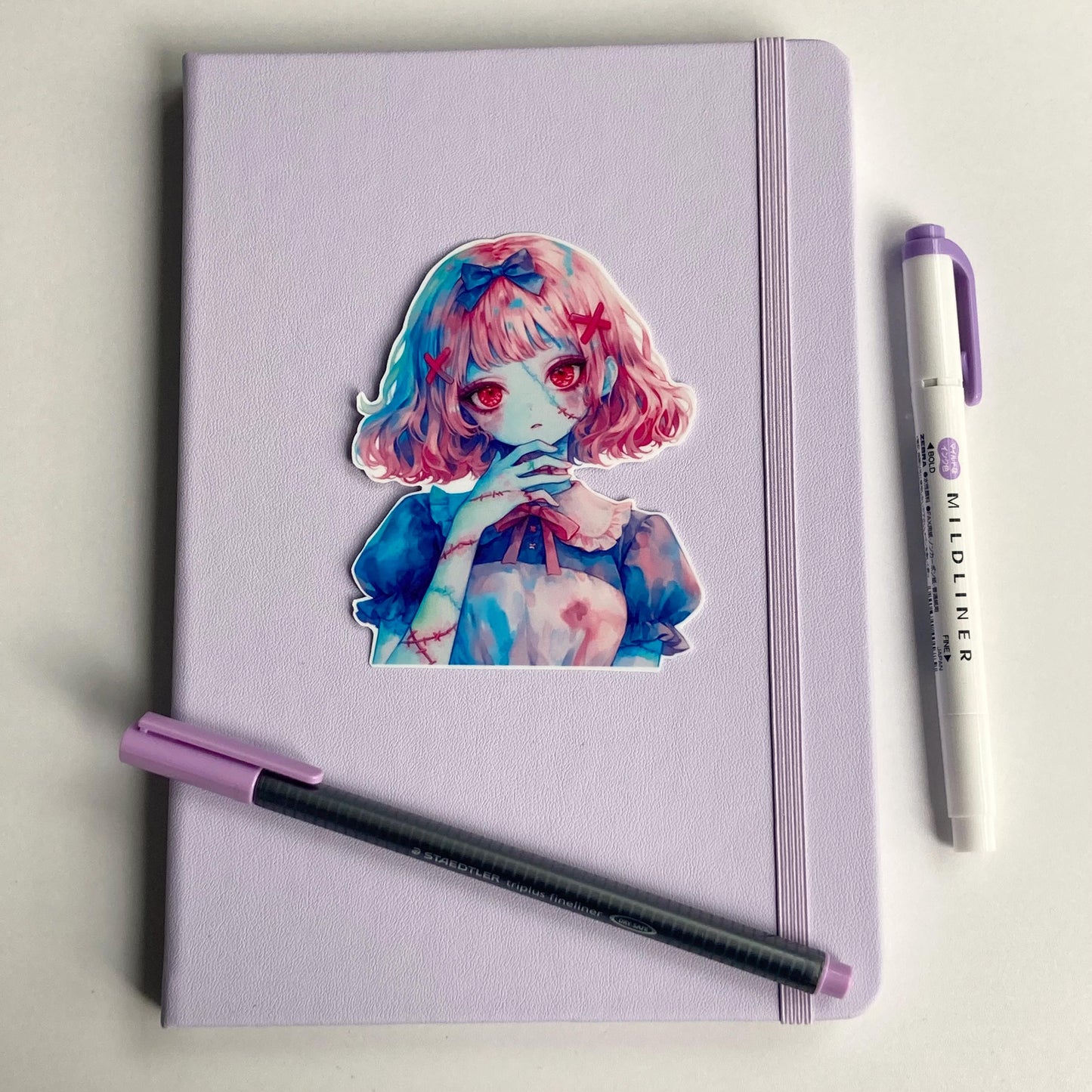 A colorful anime-style sticker of a girl with pink hair and red eyes is placed on a lavender notebook. A black pen with a purple cap and a purple highlighter are positioned beside the notebook on a white surface.