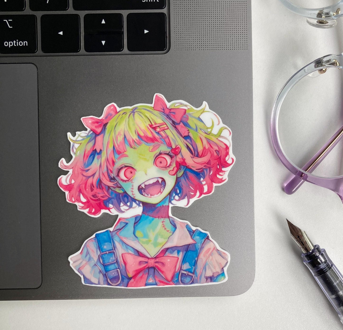 A colorful anime-style sticker of a zombie girl with green skin, pink and green hair, wide red eyes, and fangs is placed on a gray laptop. Nearby are purple-rimmed glasses and a fountain pen on a white surface.