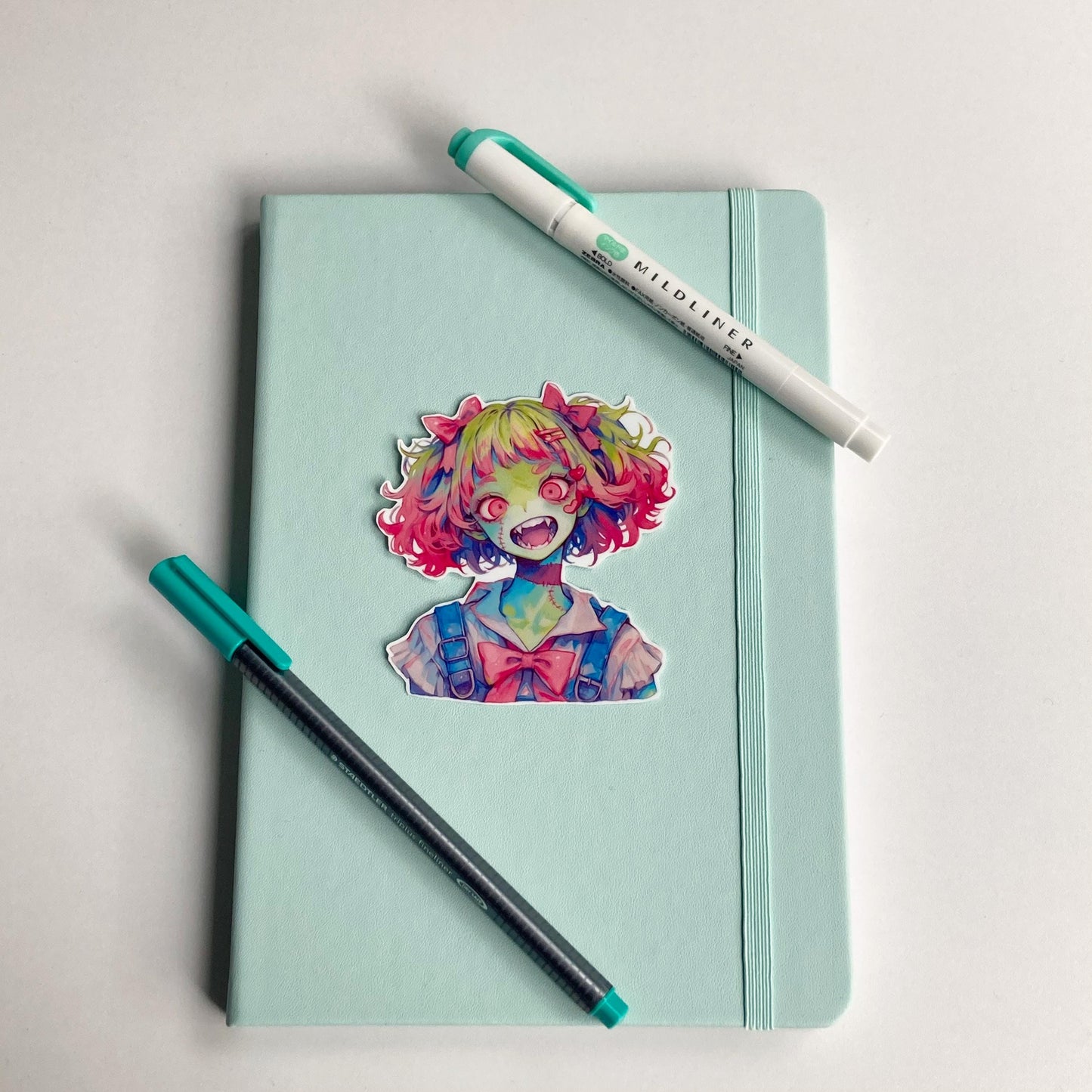 A colorful anime-style sticker of a zombie girl with green skin, pink and green hair, wide red eyes, and fangs is placed on a mint green notebook.