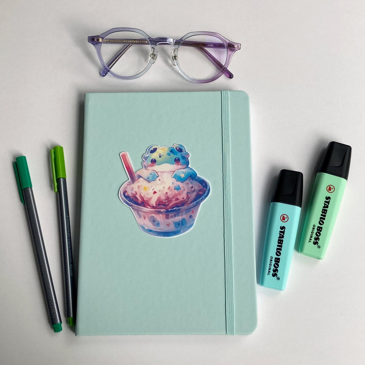 A mint green notebook with a sticker of an alien frog in a cup of pastel ice cream is surrounded by purple-rimmed glasses, two green pens, and two mint-colored highlighters. The items are arranged on a white surface.