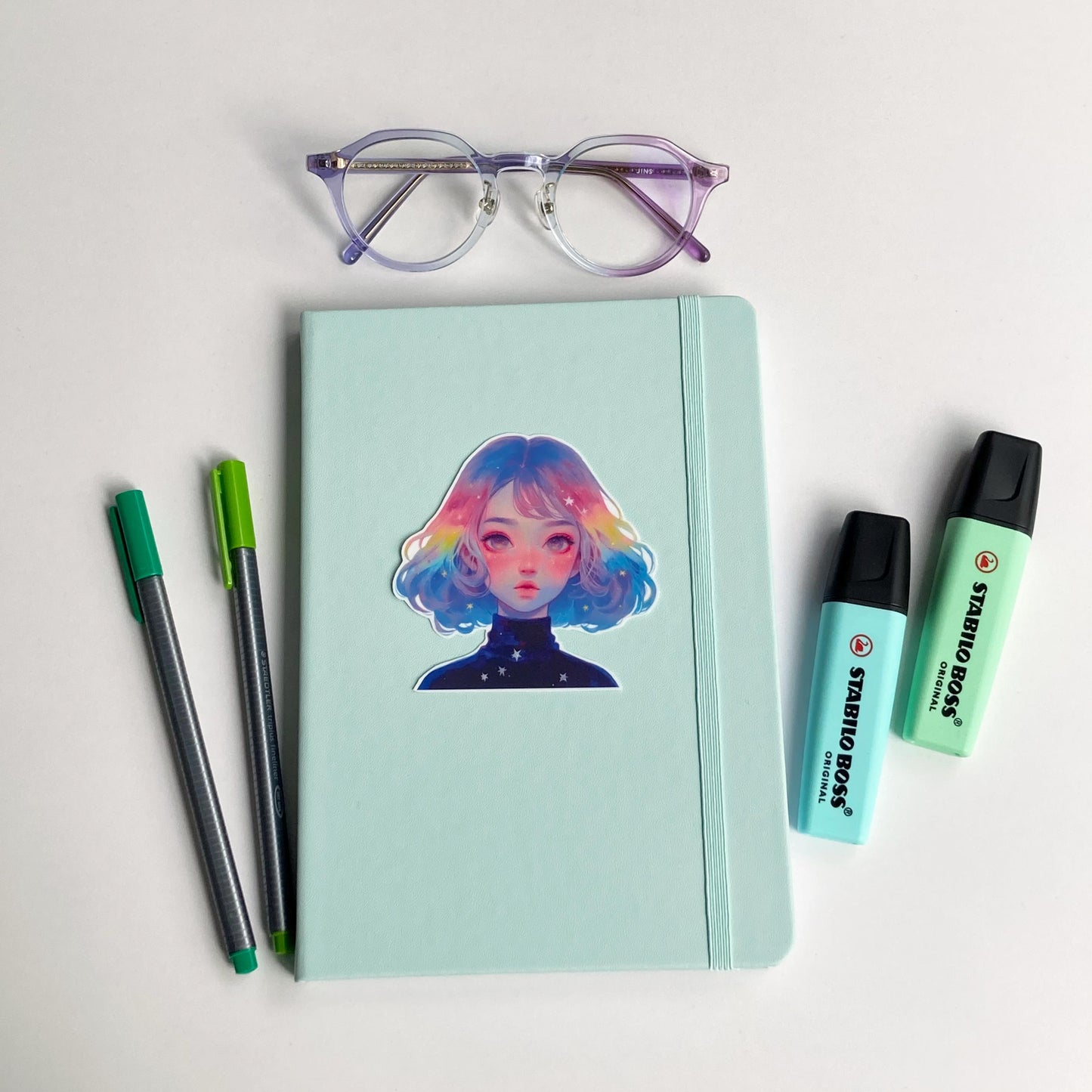 A mint green notebook with a sticker of a person with pastel rainbow hair is surrounded by purple-rimmed glasses, two green pens, and two mint-colored highlighters. The items are arranged on a white surface.