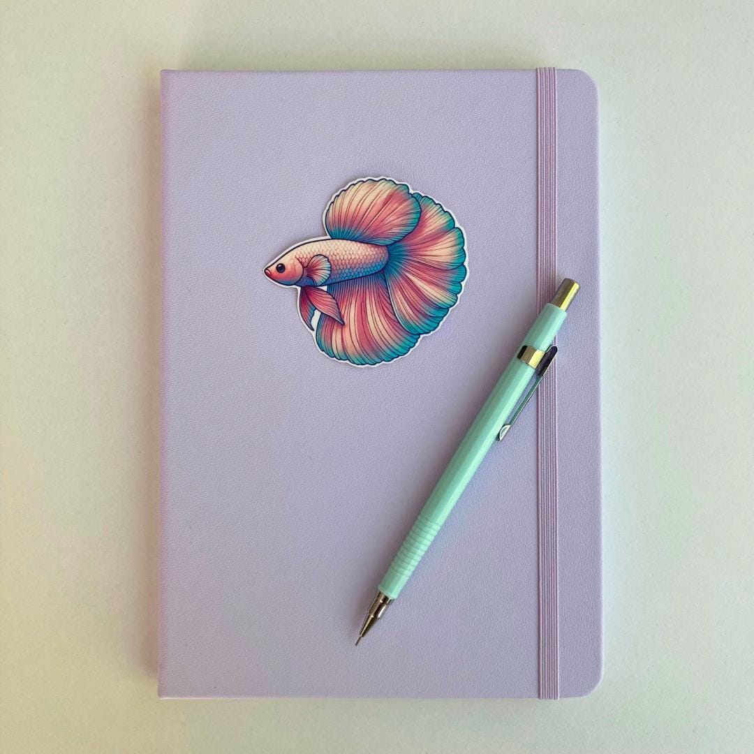A pastel purple notebook with an elastic band features a colorful betta fish sticker. A light blue mechanical pencil with gold accents rests diagonally on top. The simple background highlights the notebook and stationery items.
