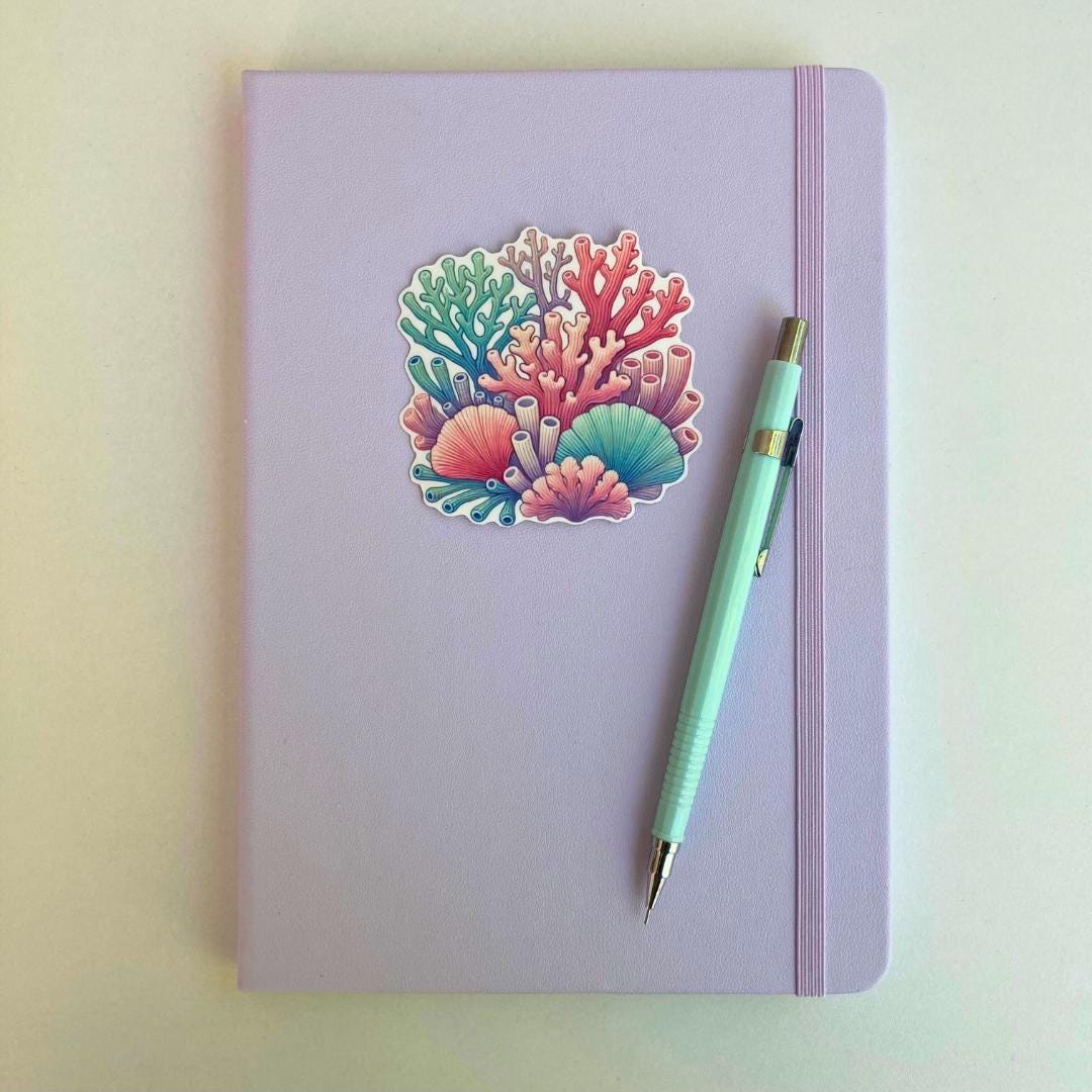 A pastel purple notebook with an elastic band features a colorful coral reef sticker in shades of pink, blue, purple, and green. A blue mechanical pencil with gold accents rests diagonally on top. The simple background highlights the stationery items