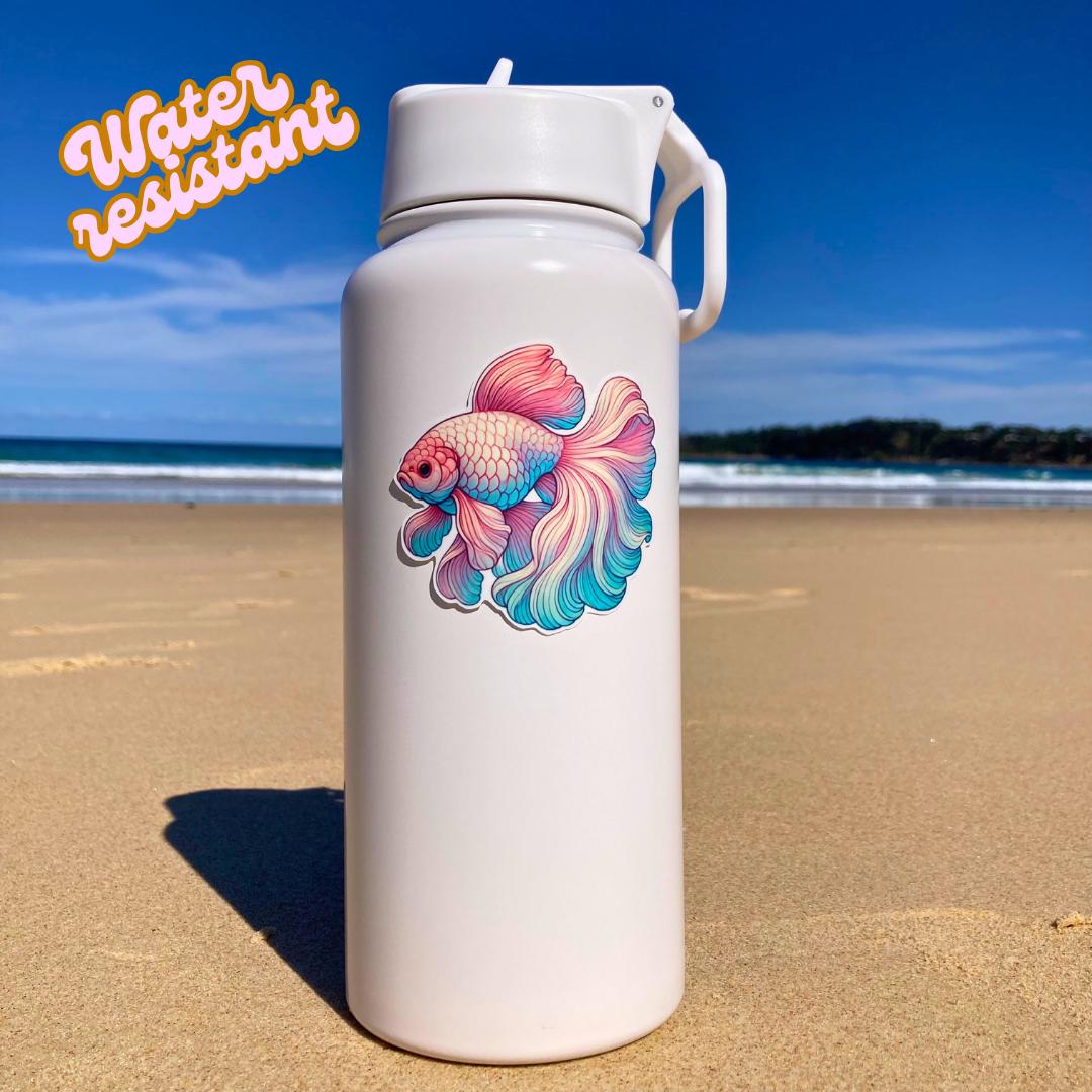 A white water bottle with a colorful betta fish sticker in pink, blue, and green hues is placed on a sandy beach. The background shows the ocean and a clear sky. The words "Water resistant" are in the top left corner, highlighting durability
