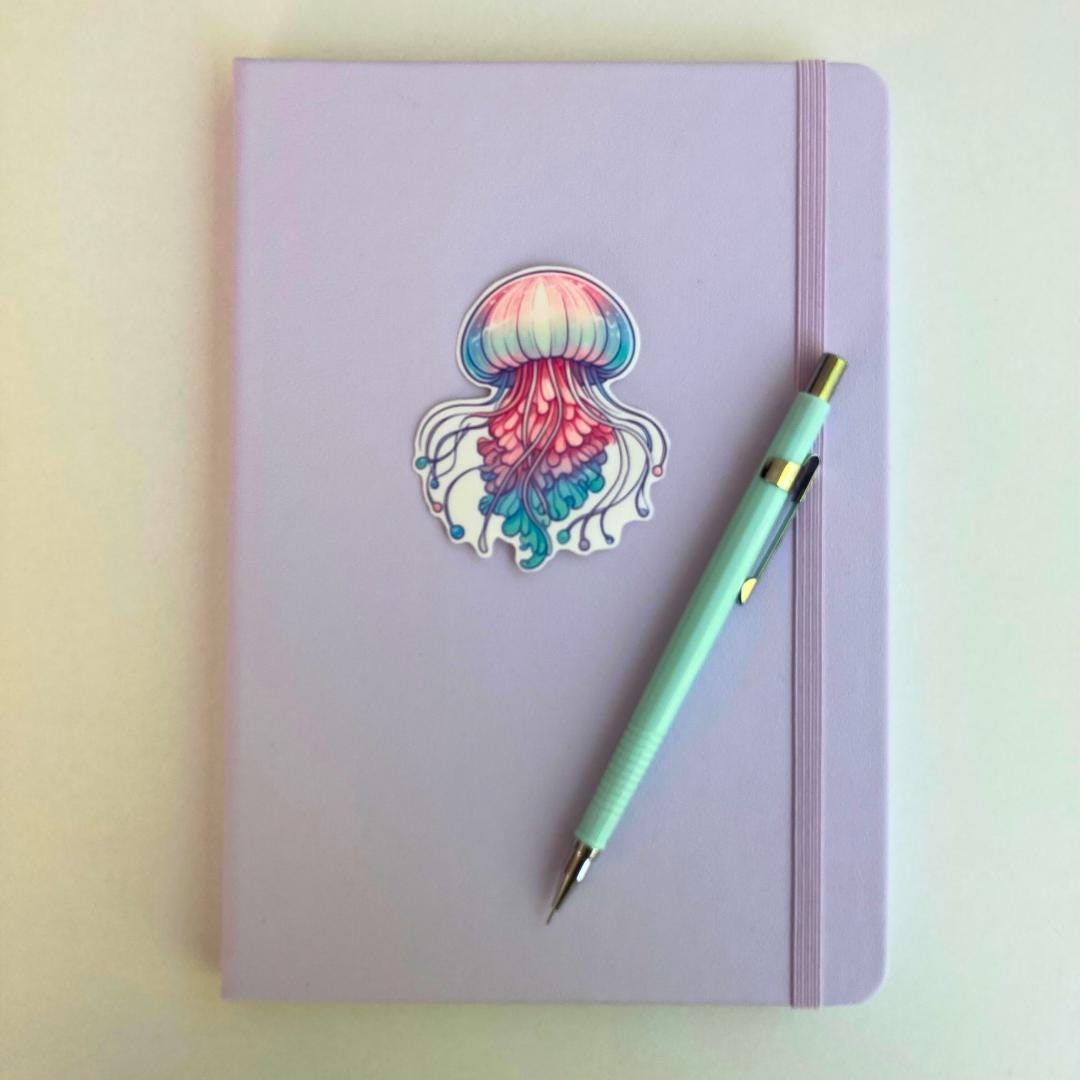 A pastel purple notebook with an elastic band features a colorful jellyfish sticker in pink, blue, and purple hues. A light blue mechanical pencil with gold accents rests diagonally on top. The simple background highlights the stationery items.