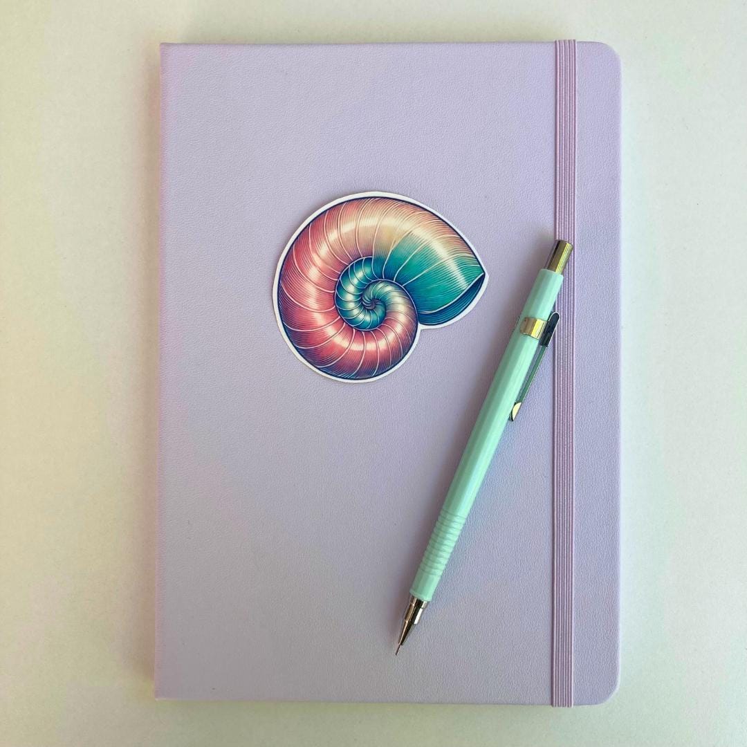 A pastel purple notebook with an elastic band features a colorful nautilus shell sticker in pink, blue, and purple hues. A light blue mechanical pencil with gold accents rests diagonally on top. The simple background highlights the stationery items.