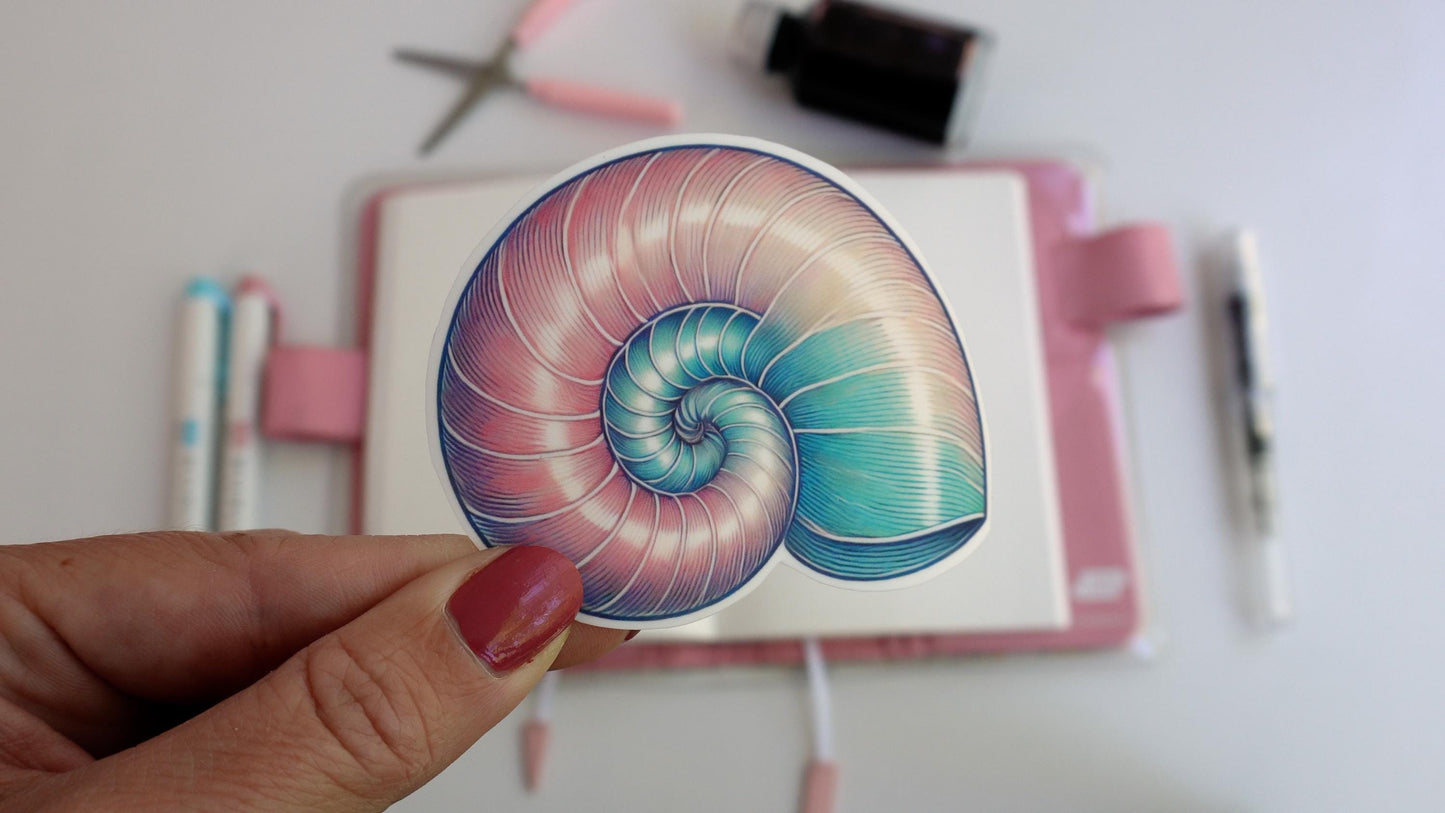 A hand holds a sticker of a pastel-colored nautilus shell with shades of pink, blue, and green. The background shows a workspace with pens, scissors, and an ink bottle on a desk, suggesting a creative setting.