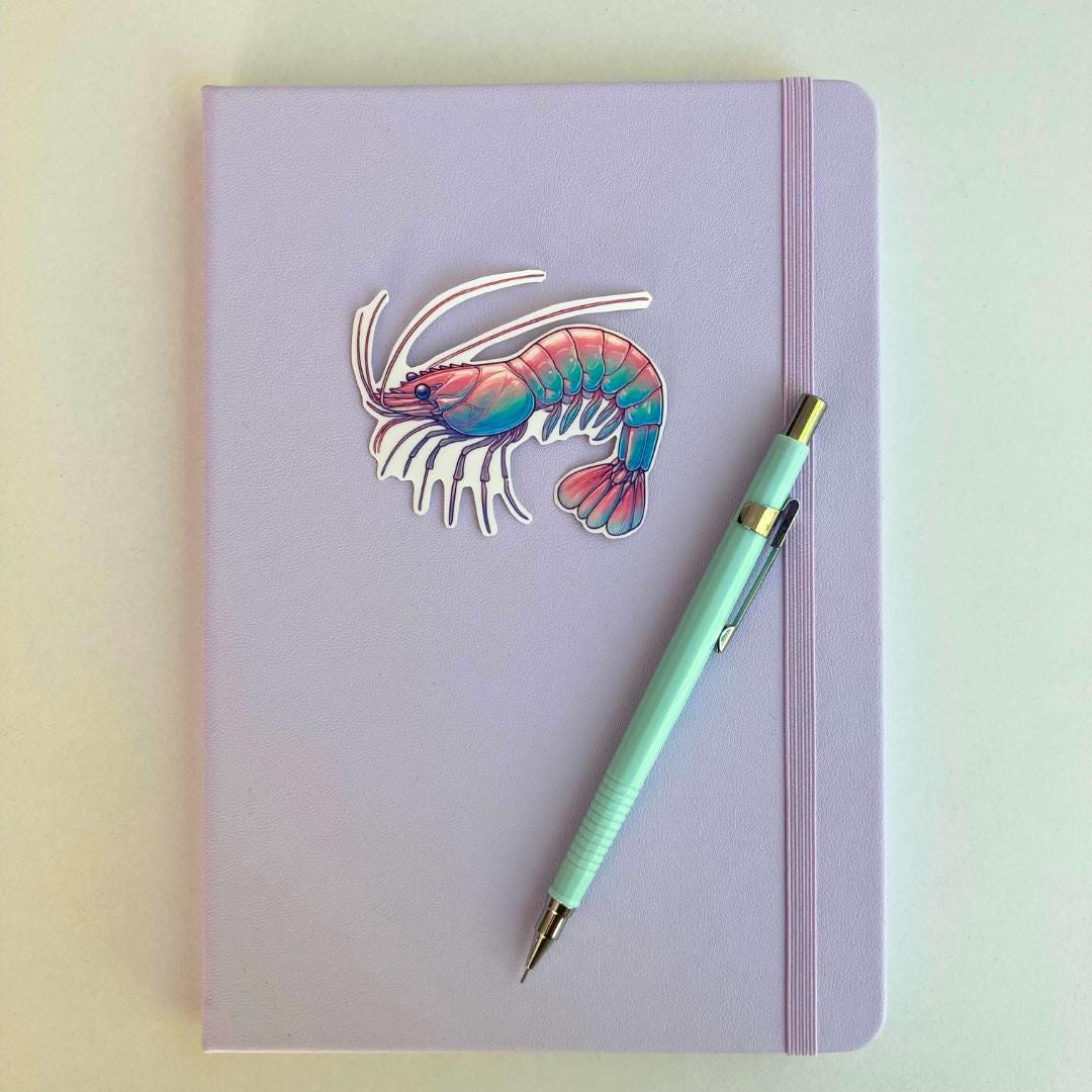 A pastel purple notebook with an elastic band features a colorful shrimp sticker in pink, blue, and purple hues. A light blue mechanical pencil with gold accents rests diagonally on top. The simple background emphasizes the stationery items.