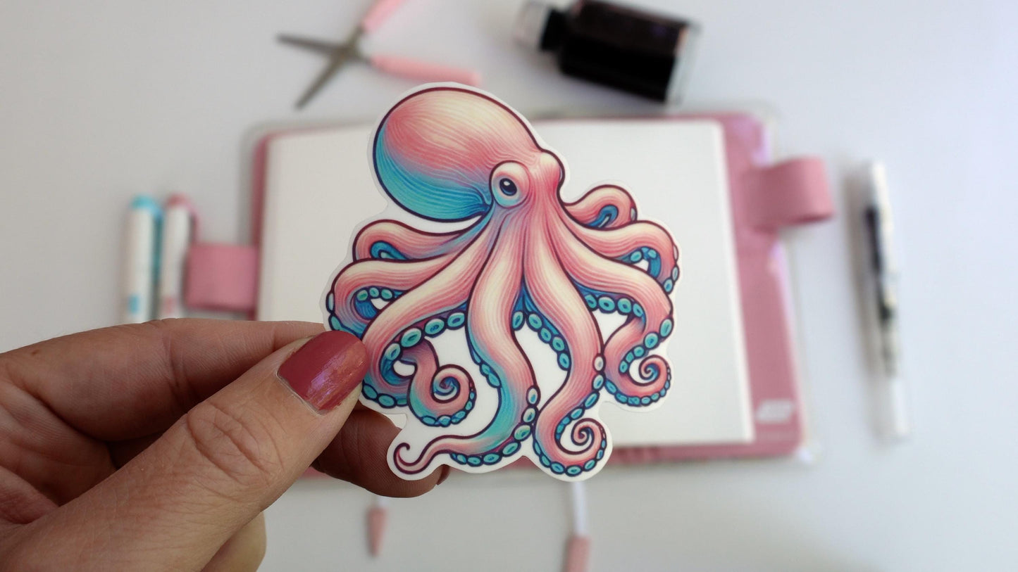 A hand holds a colorful octopus sticker in soft pink, blue, and purple tones. In the background, a desk is visible with pens, scissors, and an ink bottle, creating a creative, artistic workspace atmosphere.