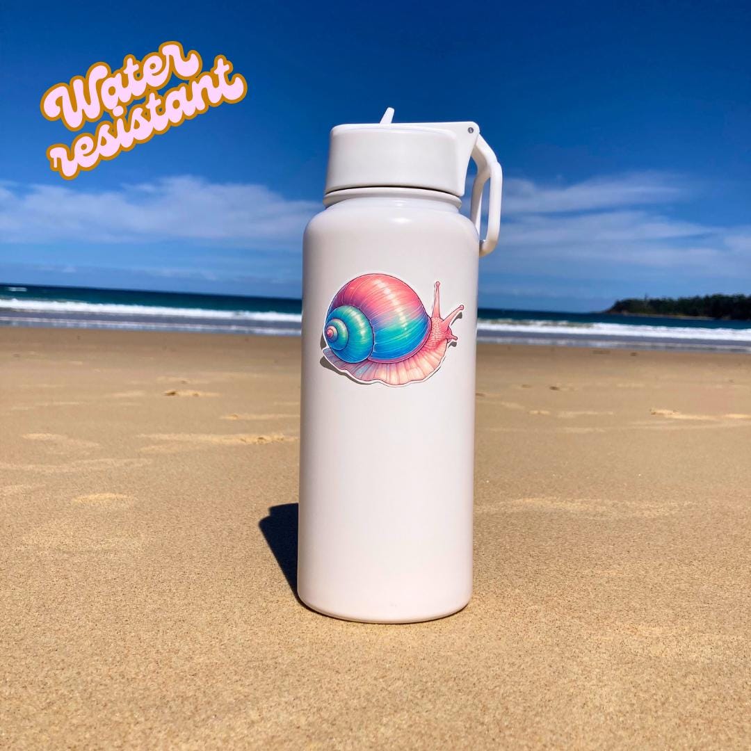 A white water bottle with a colorful snail sticker in pink, blue, and purple hues is placed on a sandy beach. The background shows the ocean and a clear sky. The words Water resistant are in the top left corner, highlighting the sticker's durability