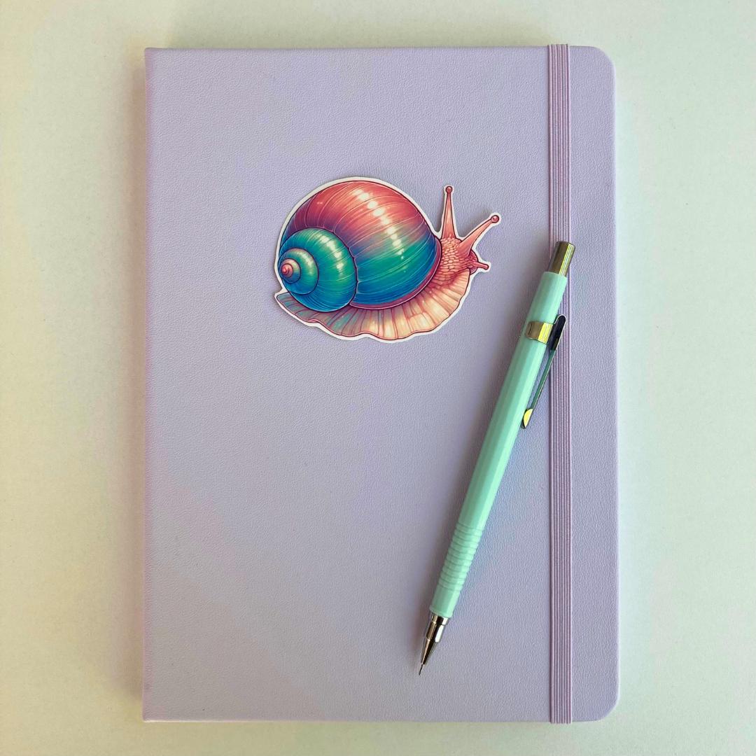 A pastel purple notebook with an elastic band features a colorful snail sticker in pink, blue, and purple hues. A light blue mechanical pencil with gold accents rests diagonally on top. The simple background highlights the stationery items.