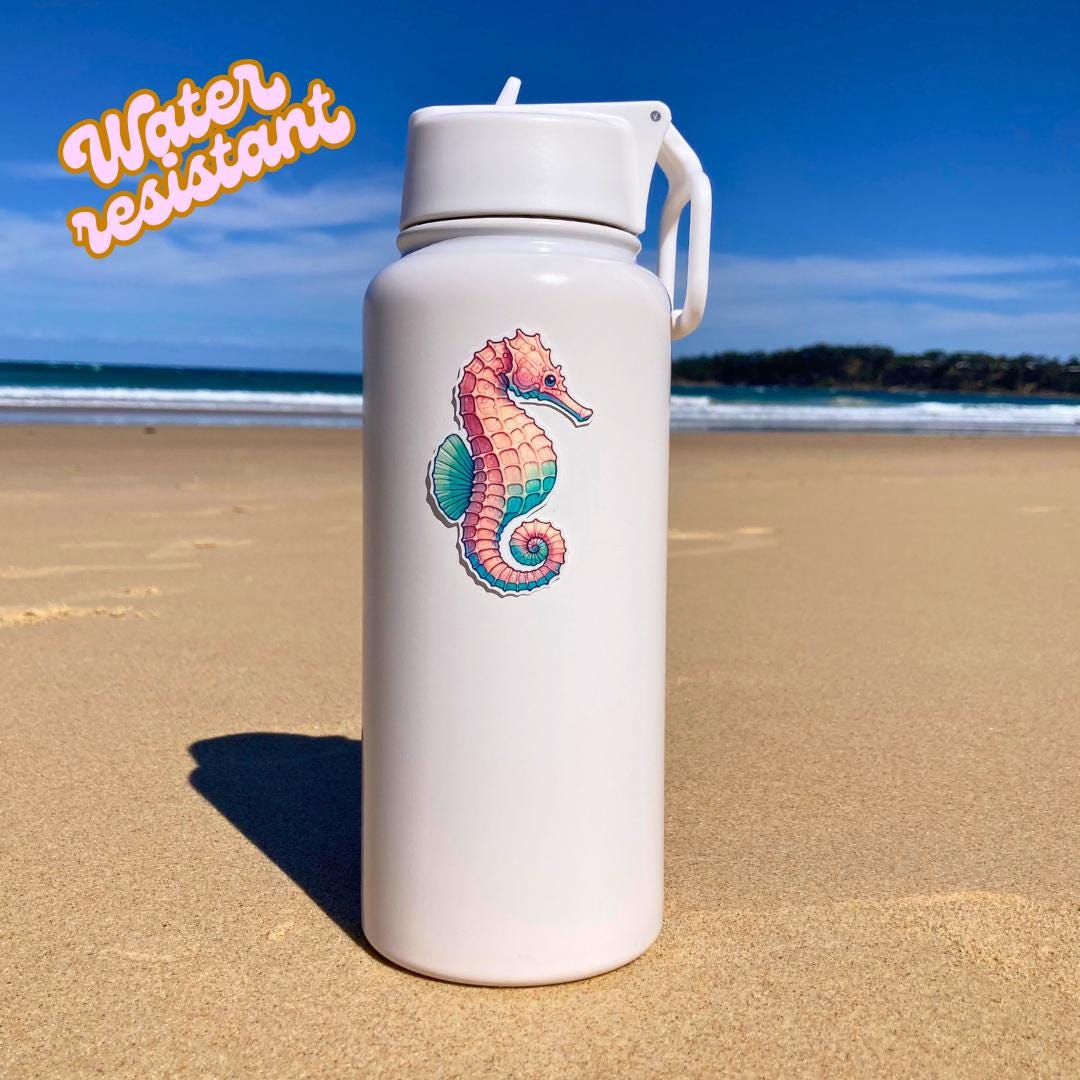 A white water bottle with a colorful seahorse sticker in pink, blue, and green hues is placed on a sandy beach. The background shows the ocean and sky. The words Water resistant are in the top left corner, highlighting the sticker's durability