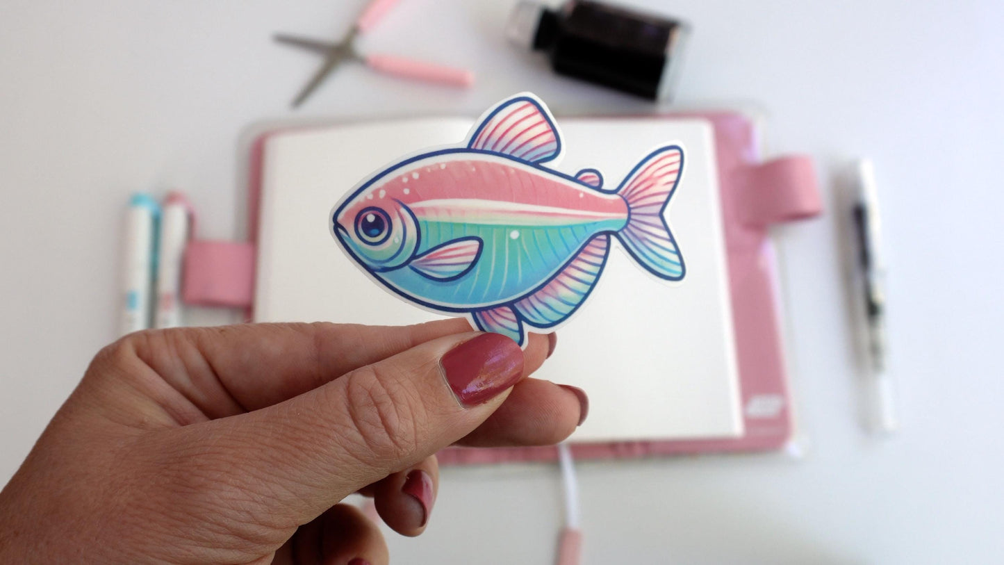 A hand holds a sticker of a pastel-colored tetra fish with pink, blue, and green hues. Behind it, a creative workspace is visible, featuring a notebook, pastel pens, scissors, and an ink bottle, all arranged on a white surface.
