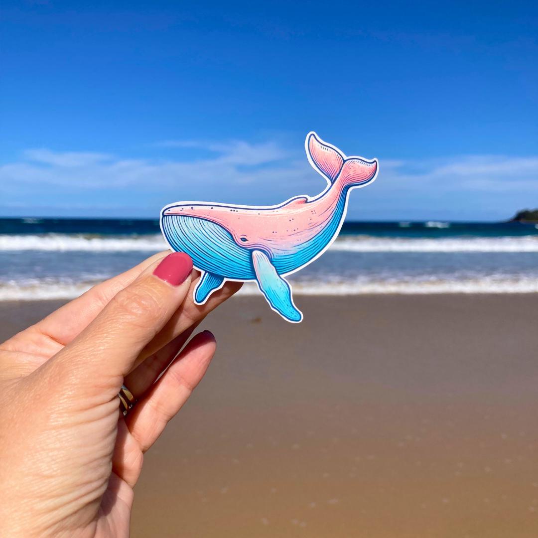 A hand with pink nails holds a colorful whale sticker with shades of pink and blue. The background shows a sandy beach with waves and a clear blue sky, highlighting the whale's playful design against the serene coastal setting.