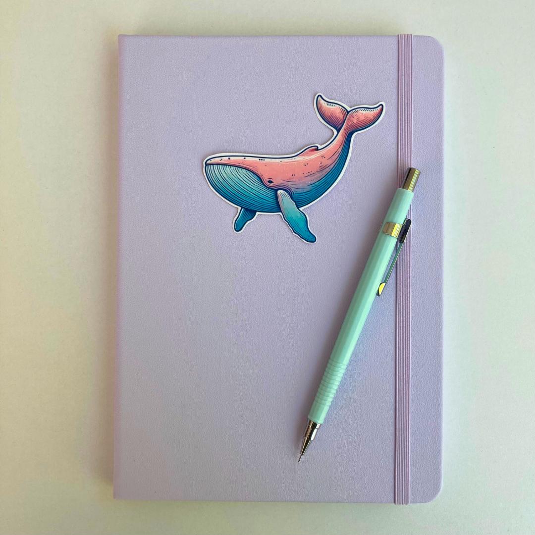 A pastel purple notebook with an elastic band features a colorful whale sticker in pink and blue hues. A light blue mechanical pencil with gold accents rests diagonally on top. The simple background highlights the notebook and stationery items.