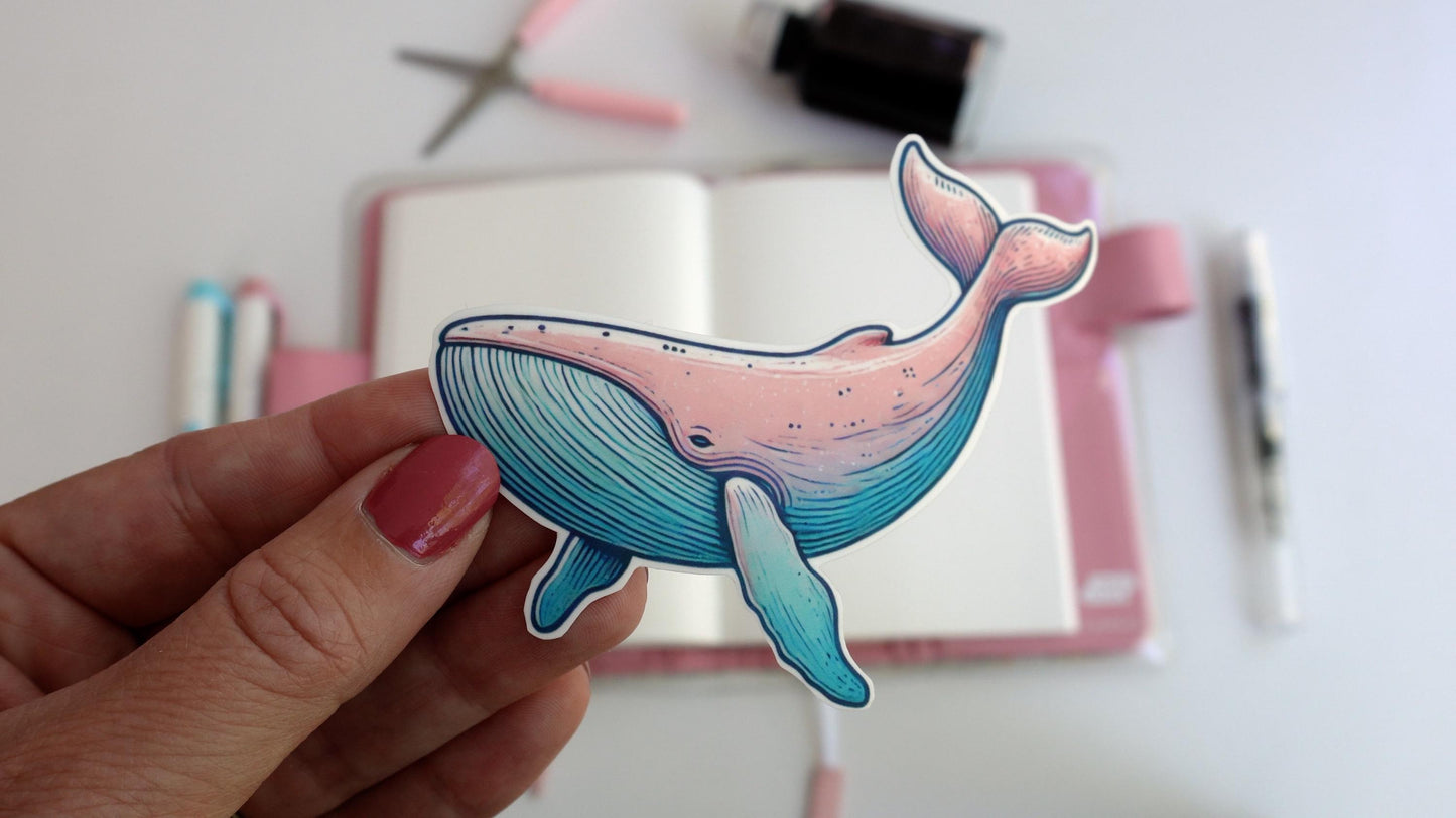 A hand holds a pastel-colored whale sticker, blending shades of pink and blue. The background features a creative workspace with an open notebook, pastel pens, scissors, and an ink bottle arranged on a white desk.