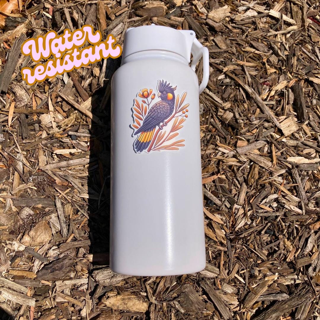 A white water bottle with a sticker of a black cockatoo perched on a branch with orange flowers and leaves lies on wood chips in bright sunlight. An orange "Water resistant" label is in the top left corner.