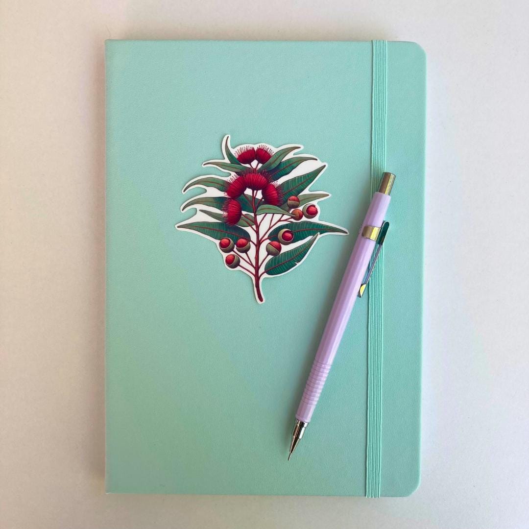 A light turquoise notebook with an elastic strap features a sticker of a eucalyptus branch with red blossoms and buds. A lavender mechanical pencil is placed diagonally on the notebook, which lies on a white surface.