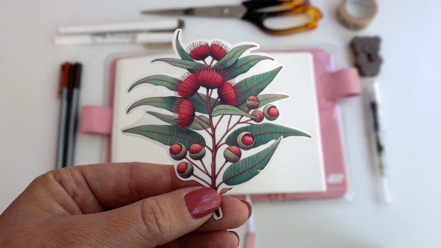 A hand with pink nail polish holds a sticker of a eucalyptus branch with red blossoms and buds. In the background, an open notebook with a pink cover sits on a white surface along with scattered pens, scissors, and a roll of tape
