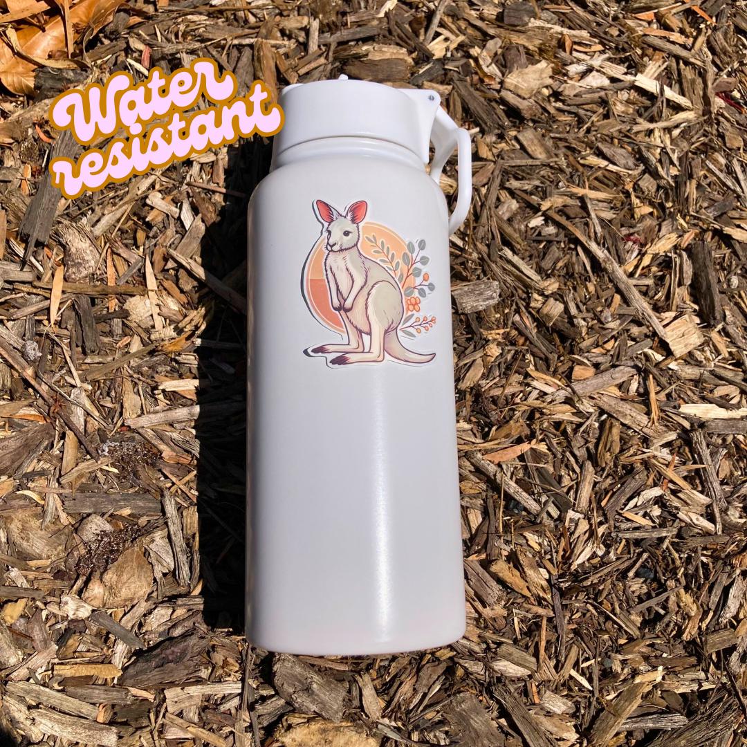 A white water bottle with a cartoon kangaroo sticker is placed on wood chips in bright sunlight. The sticker shows a white kangaroo with a tan circle and leaves in the background. An orange "Water resistant" label is in the top left corner.