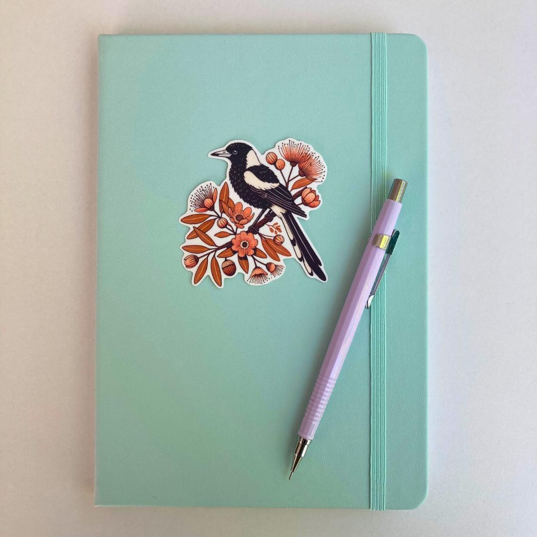 A light turquoise notebook with an elastic strap features a sticker of a black-and-white magpie perched on a branch with orange flowers and leaves. A lavender mechanical pencil is placed diagonally on the notebook, which rests on a white surface.