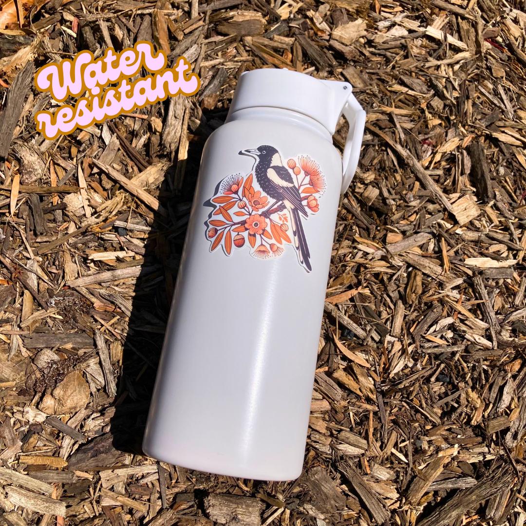 A white water bottle with a sticker of a black-and-white magpie perched on a branch with orange flowers and leaves lies on wood chips in bright sunlight. An orange "Water resistant" label is in the top left corner.