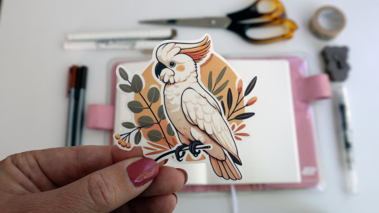 A hand holds a white cockatoo sticker featuring an orange crest. The bird perches on a branch, surrounded by leaves and small orange flowers. The background is light orange. Desk tools like pens and scissors are blurred in the background.
