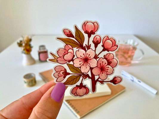 A hand with purple-painted nails holds a cartoon-style cherry blossom sticker, featuring pink blooms with red accents and brown stems. The background shows a white table with dried flowers, a tea cup, a notebook, and a pen.