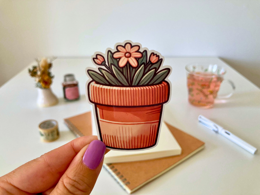 A hand with purple nail polish holds a cartoon sticker of a potted plant with orange flowers. The background features a desk with a notebook, pen, dried flowers, a jar, and a glass cup of tea.