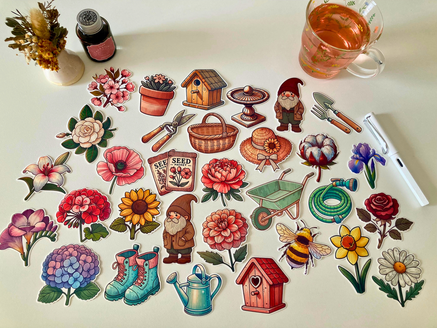 A collection of garden-themed stickers, including flowers, gardening tools, seed packets, gnomes, a birdhouse, boots, a watering can, a wheelbarrow, and a bee. Nearby are dried flowers, a tea cup, a white pen, and an ink bottle on a white table.