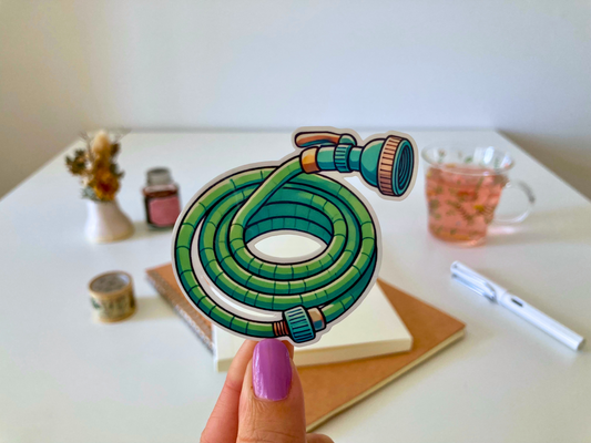 A hand with purple-painted nails holds a cartoon-style garden hose sticker, coiled and green with an orange nozzle. The background shows a white table with dried flowers, a tea cup, a notebook, and a pen.