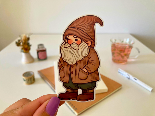 A hand holds a sticker of a gnome wearing a brown hat and coat, with a white beard and rosy cheeks. The background shows a desk with notebooks, dried flowers, a pen, and a cup of tea.