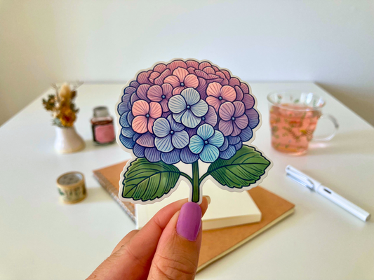 A hand with purple-painted nails holds a cartoon-style hydrangea sticker, featuring pink, purple, and blue blooms with green leaves. The background shows a white table with dried flowers, a tea cup, a notebook, and a pen.