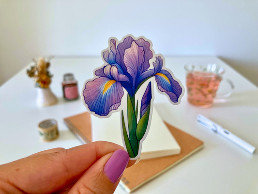 A hand with purple-painted nails holds a cartoon-style iris flower sticker, featuring purple petals with yellow accents and green leaves. The background shows a white table with dried flowers, a tea cup, a notebook, and a pen.