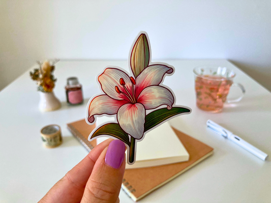 A hand with purple-painted nails holds a cartoon-style lily sticker, featuring white petals with pink accents and green leaves. The background shows a white table with dried flowers, a tea cup, a notebook, and a pen.