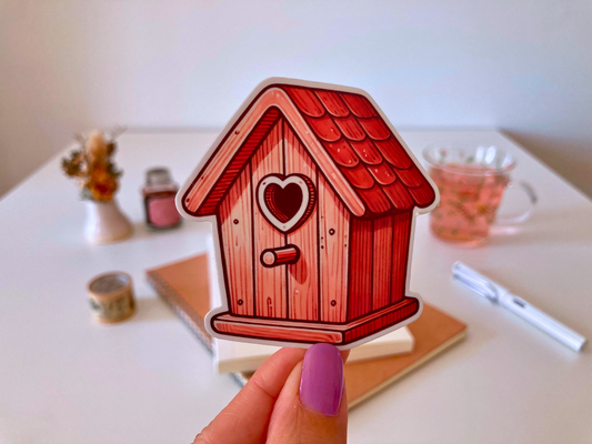 A hand with purple nails holds a birdhouse sticker with a red roof, wooden texture, and a heart-shaped entrance. The background shows a desk with a notebook, dried flowers, a tea cup, and stationery.