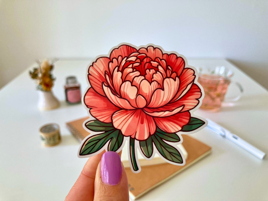 A hand with purple-painted nails holds a cartoon-style peony sticker, featuring layered red and pink petals with green leaves. The background shows a white table with dried flowers, a tea cup, a notebook, and a pen.