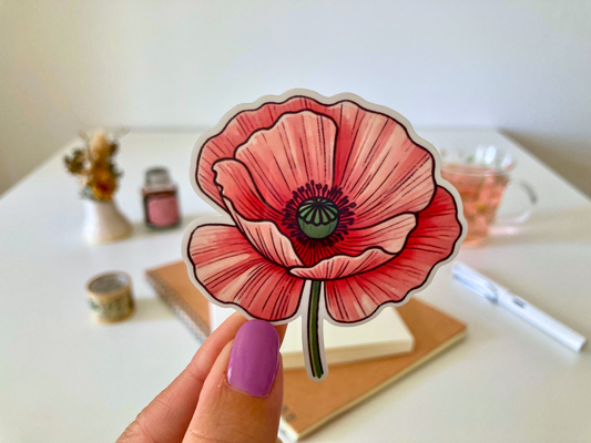 A hand with purple-painted nails holds a cartoon-style pink poppy sticker, featuring layered pink petals, a green center, and black detailing. The background shows a white table with dried flowers, a tea cup, a notebook, and a pen.