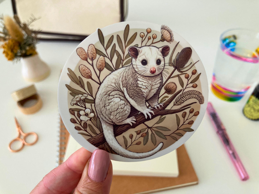 A hand with pink nails holds a round Australian possum sticker featuring a fluffy possum perched on a branch, surrounded by native botanical elements in warm earthy tones. The background includes a cozy desk setup with stationery, a notebook, a pink pen, a rainbow-rimmed glass, and dried flowers in a vase. The waterproof and scratch-resistant vinyl sticker is perfect for decorating water bottles, laptops, and journals with a touch of Australian wildlife charm.