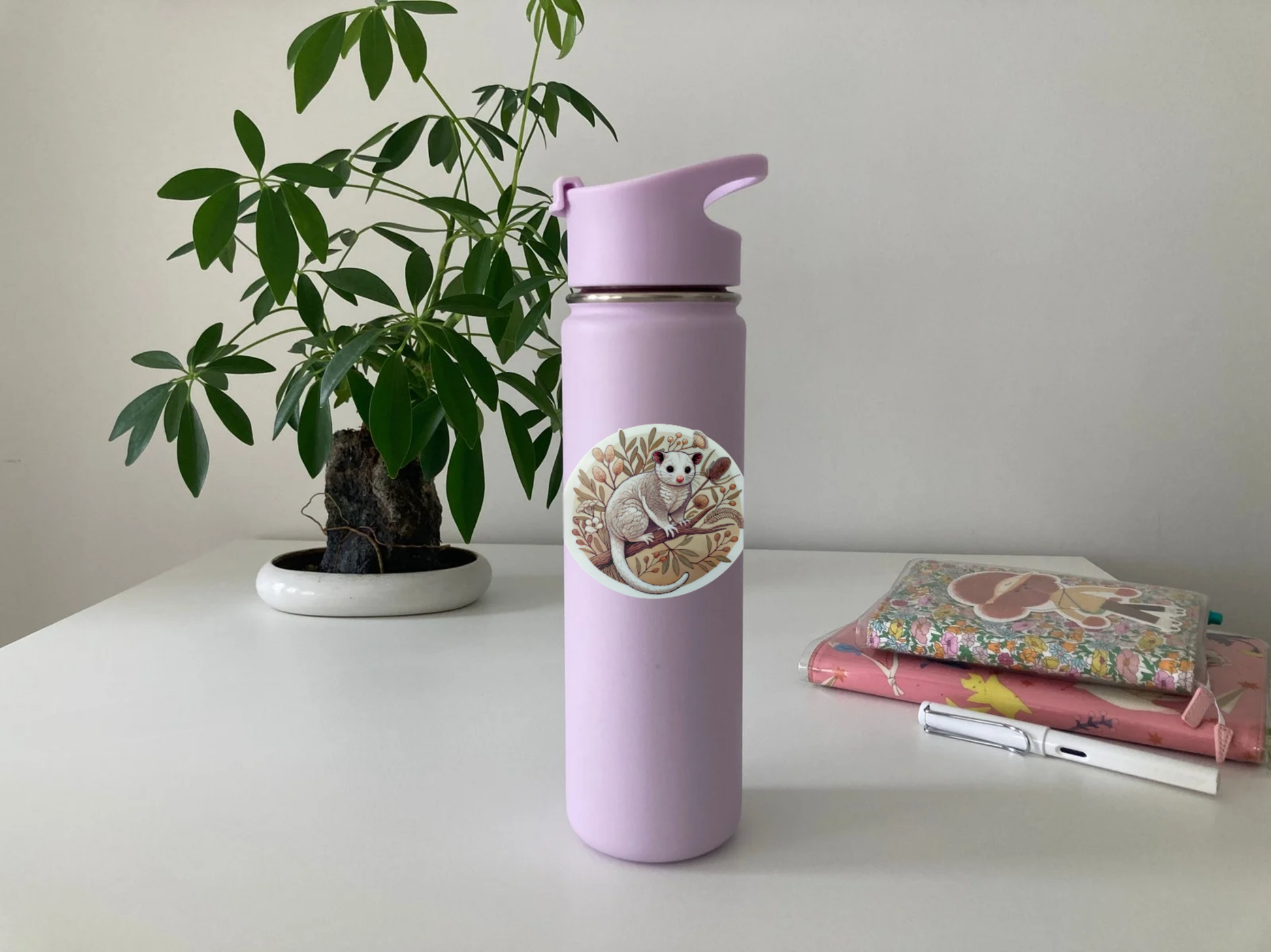 A lavender drink bottle with a round Australian possum sticker sits on a white table. The sticker features a fluffy possum perched on a branch, surrounded by native botanicals in warm earthy tones. In the background, a bonsai-style plant in a white pot, a stack of pastel notebooks with stickers, and a white fountain pen add a cozy touch. The waterproof, scratch-resistant sticker is perfect for decorating bottles, laptops, and journals with a nature-inspired aesthetic.