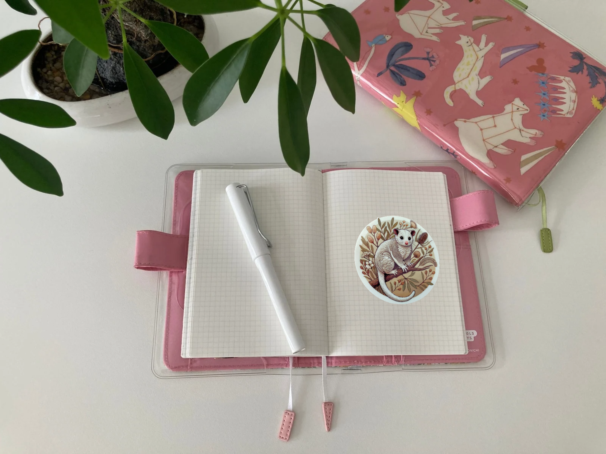 An open Hobonichi Techo journal with a pink cover lies on a white desk, displaying a round Australian possum sticker on its grid paper pages. The sticker features a fluffy possum perched on a branch, surrounded by native botanicals in warm earthy tones. A white fountain pen rests on the journal, and a pink animal-themed notebook sits nearby. Green plant leaves frame the top of the image, adding a cozy, nature-inspired touch to the workspace.