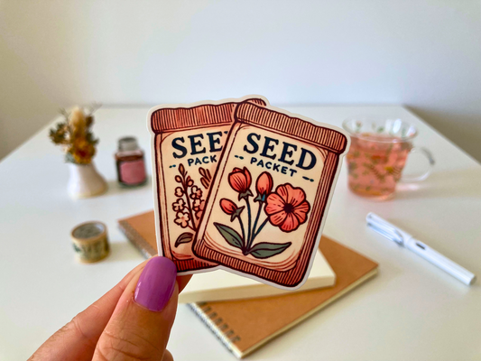 A hand with purple nail polish holds two cartoon seed packet stickers featuring illustrated flowers. The background shows a desk with a notebook, pen, dried flowers, washi tape, a jar, and a glass cup of tea.